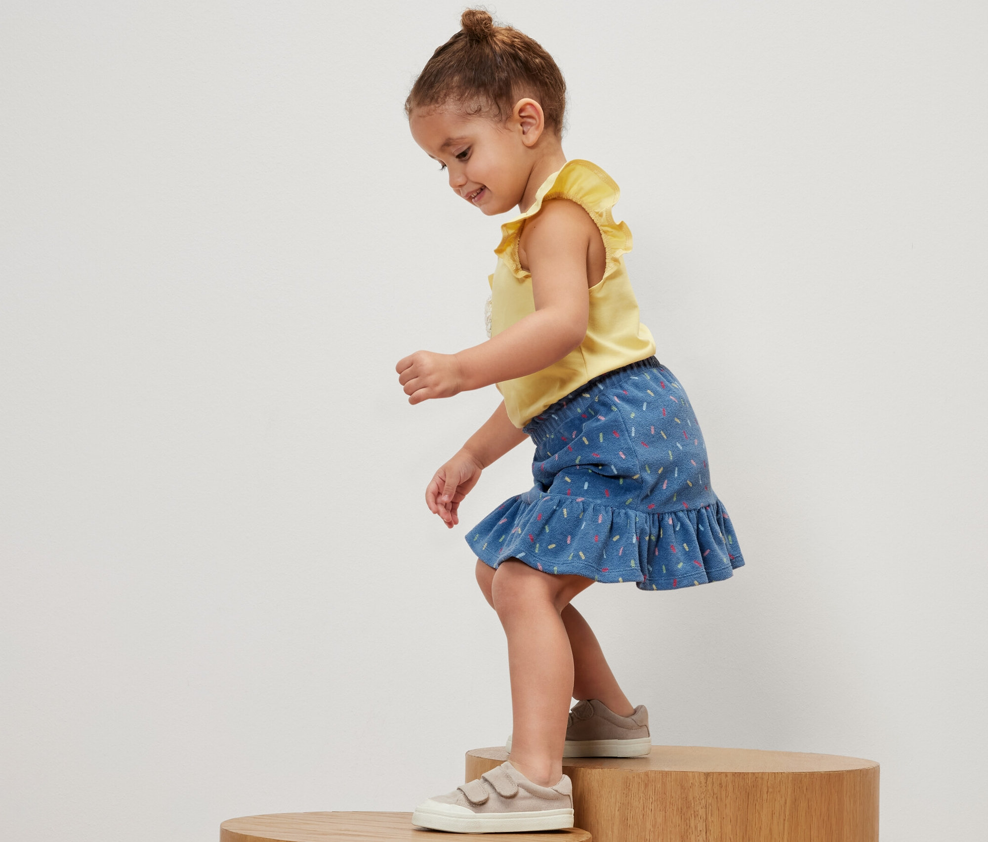 Dievčatko stojí na drevenom podstavci a má na sebe žlté tričko a dievčenskú froté sukňu.