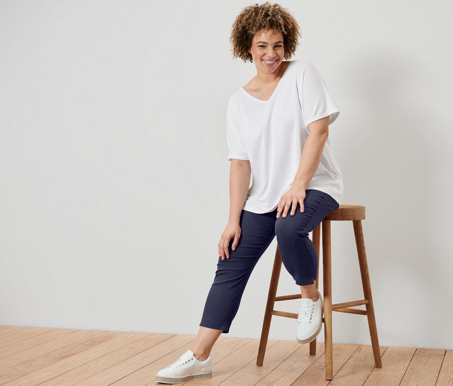 Žena sedí na stoličke a má na sebe biele štruktúrované tričko a tmavomodré sedemosminové elastické nohavice.