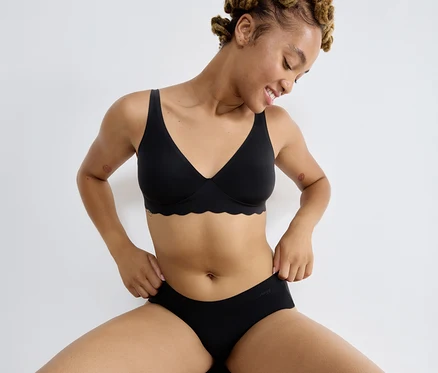 Usmievavá žena sedí na stoličke, oblečená v čiernej Podprsenke sloggi ZERO Microfibre 2.0 Soft bra a čiernych nohavičkách.