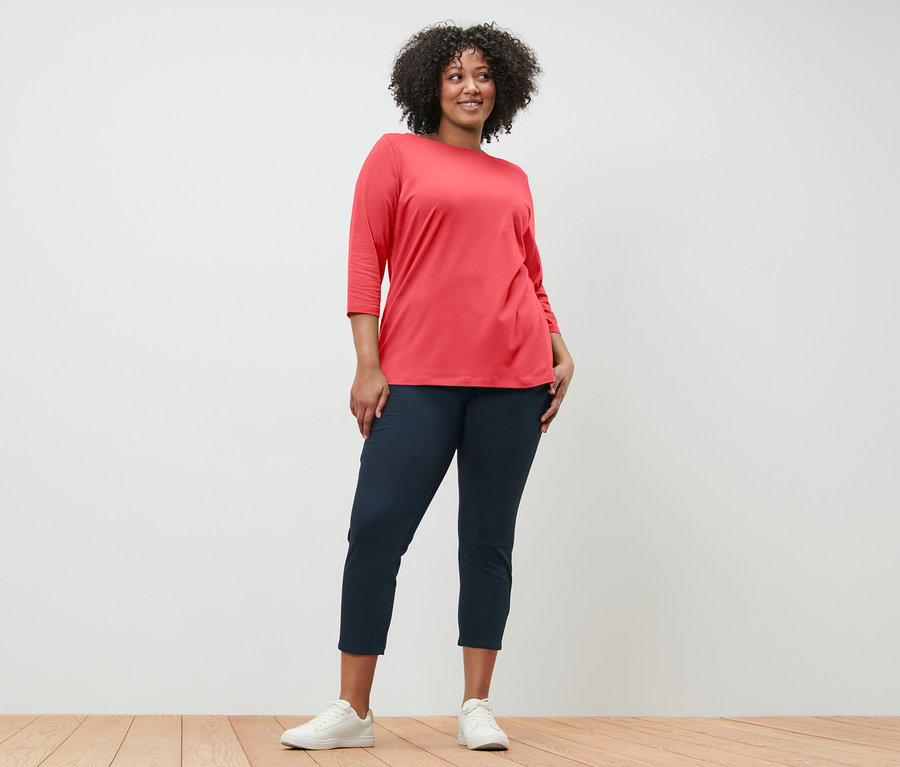 Žena pózuje v koralovom tričku s trojštvrťovým rukávom a tmavomodrých nohaviciach.