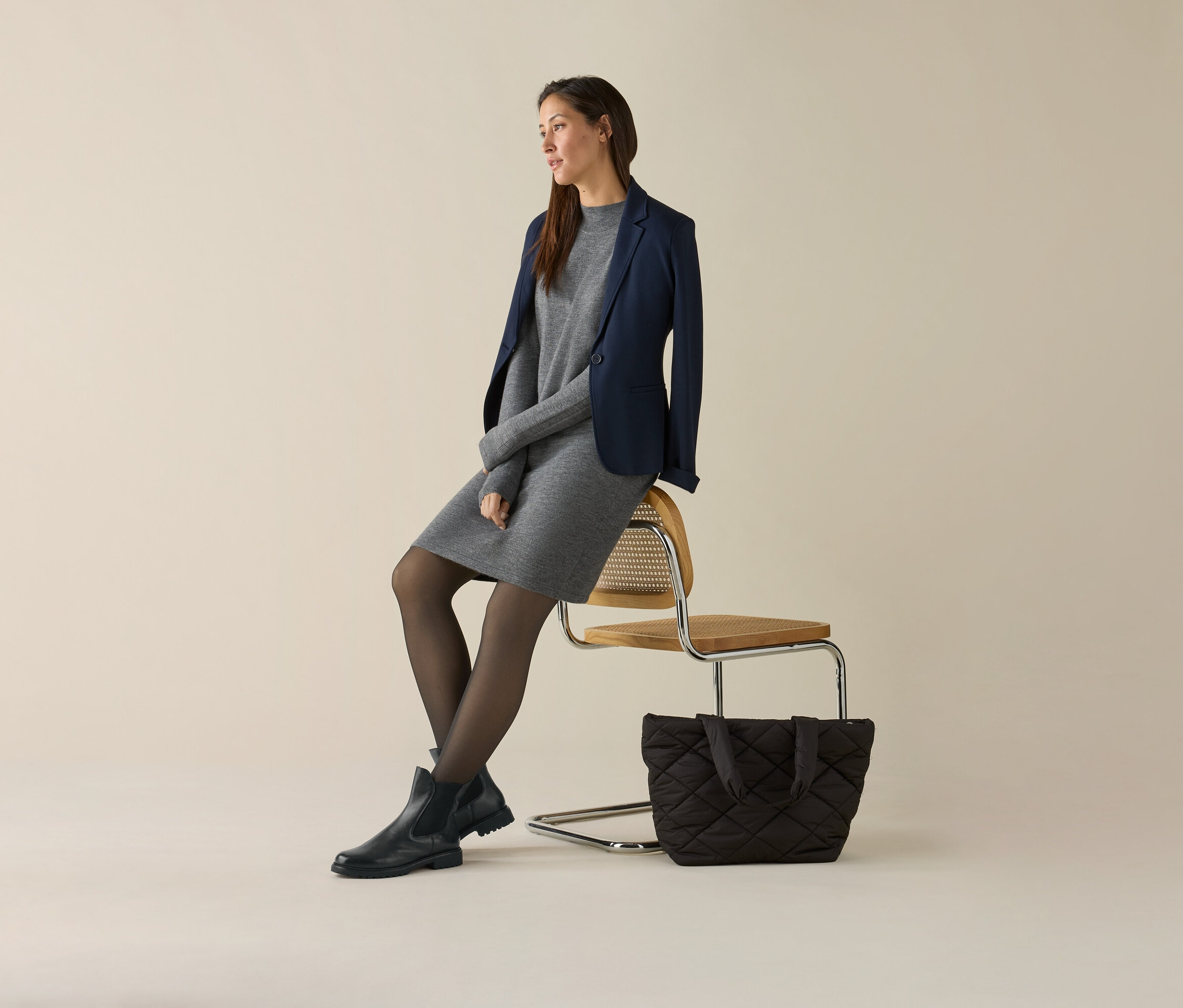Žena sedí na stoličke v sivých šatách a tmavomodrom džersejovom saku. Vedľa stoličky je kabelka.
