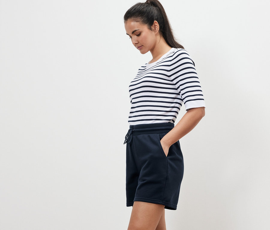 Žena v pruhovanom tričku a tmavomodrých teplákových šortkách stojí bokom.