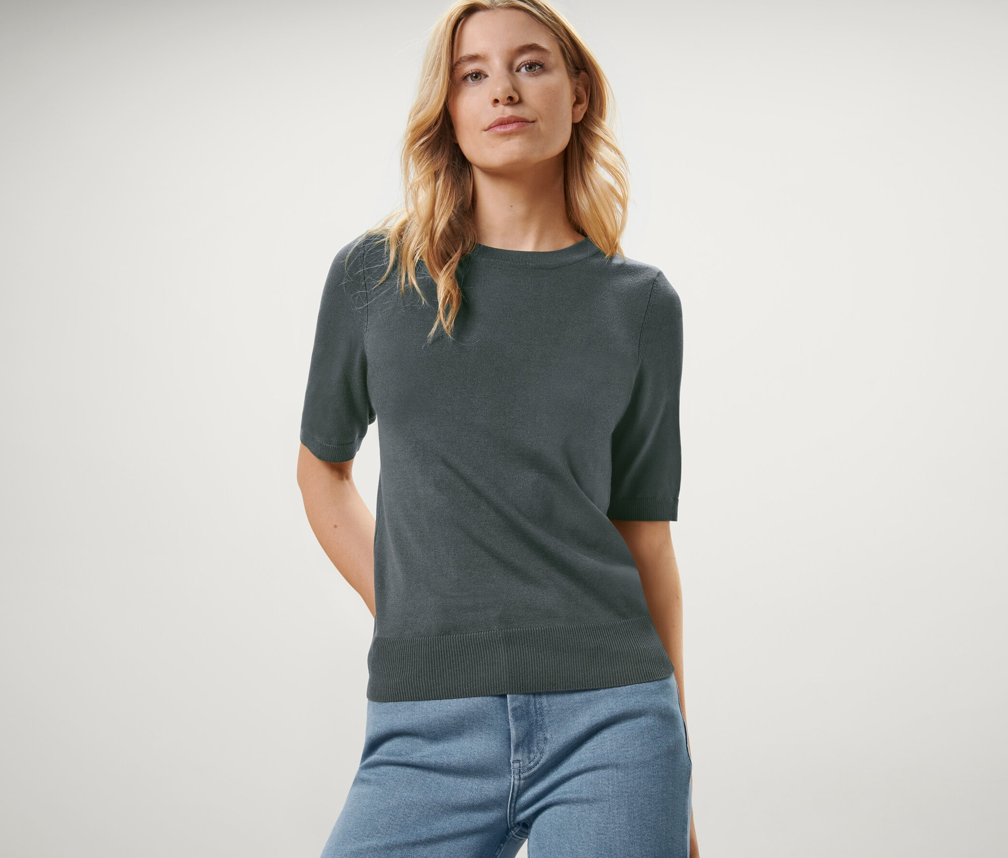 Žena pózuje v zelenom pulóvri z jemného úpletu a modrých džínsoch.