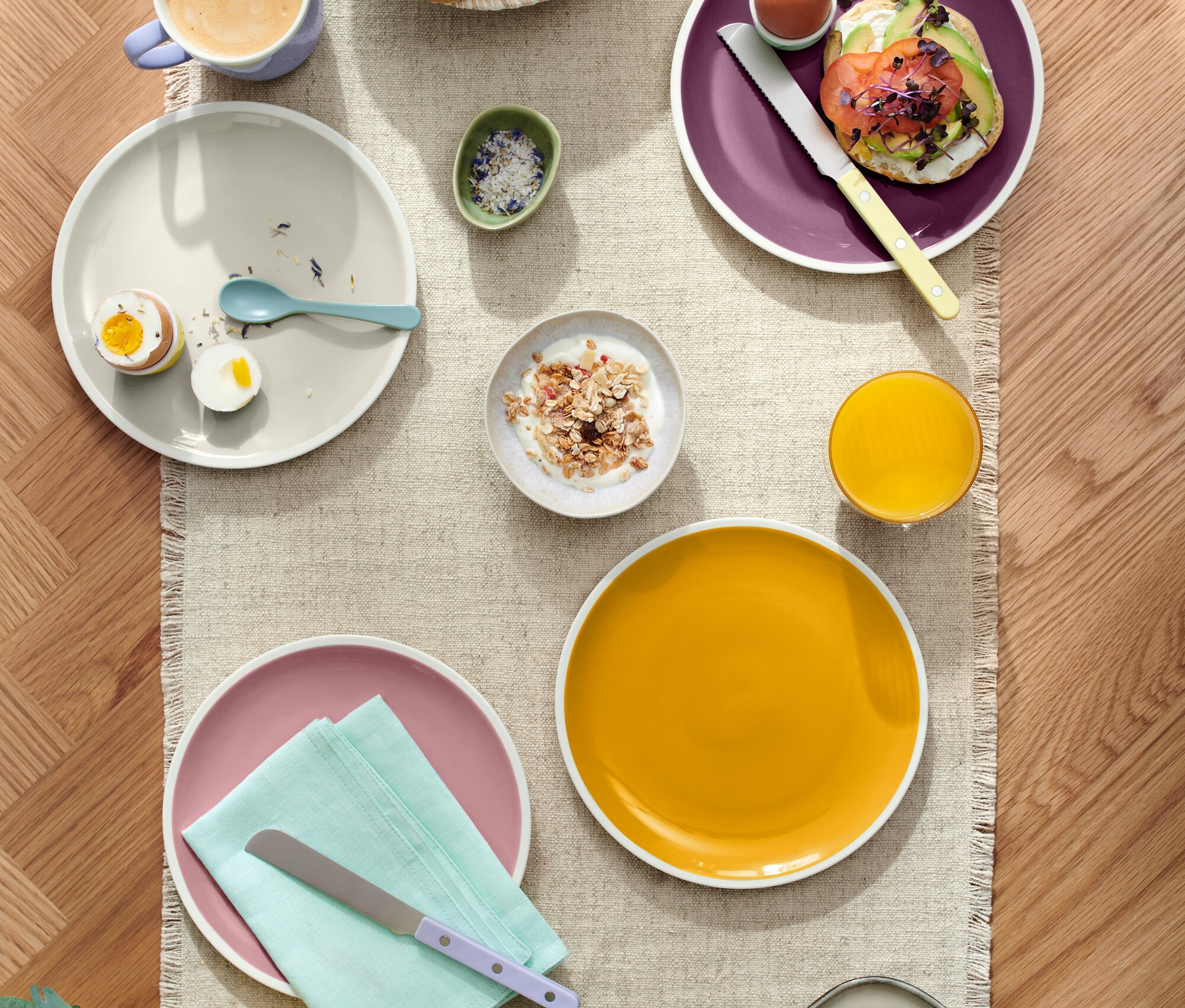 Pohľad zhora na prestretý stôl s rôznymi jedlami, vrátane pečiva v košíku, tanierov s vajíčkami, avokádovým toastom a syrom, spolu s nápojmi ako káva a džús.