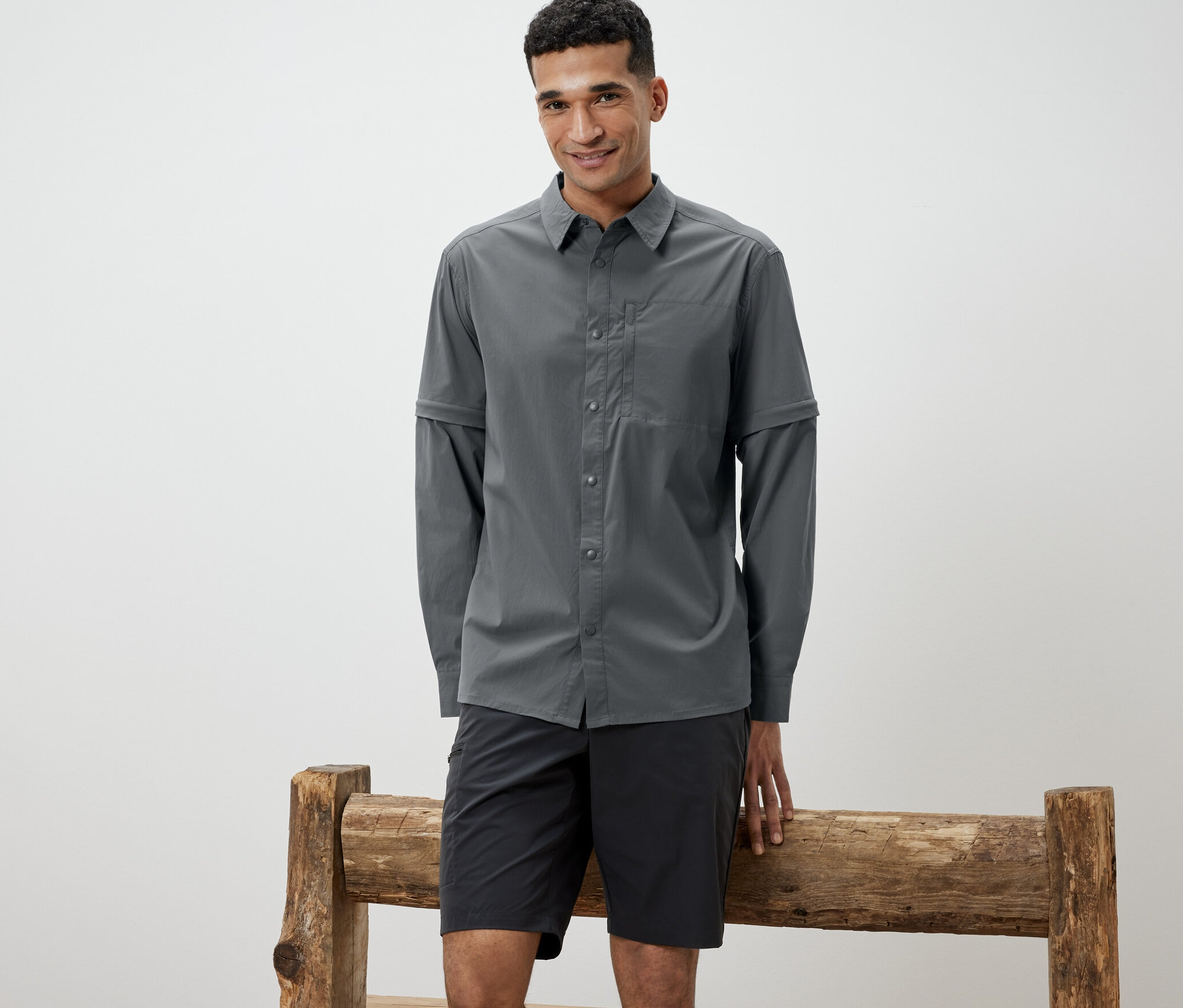 Muž opretý o drevenú lavičku má na sebe sivú outdoorovú košeľu a čierne šortky.