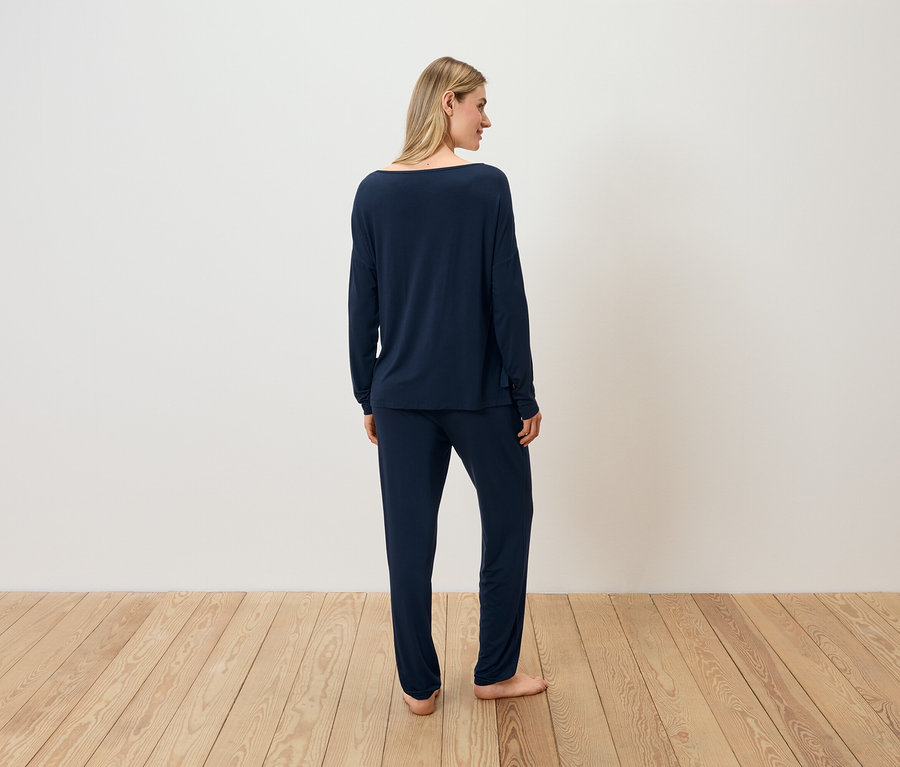 Žena v tmavomodrom pyžame stojí chrbtom ku kamere na drevenej podlahe.