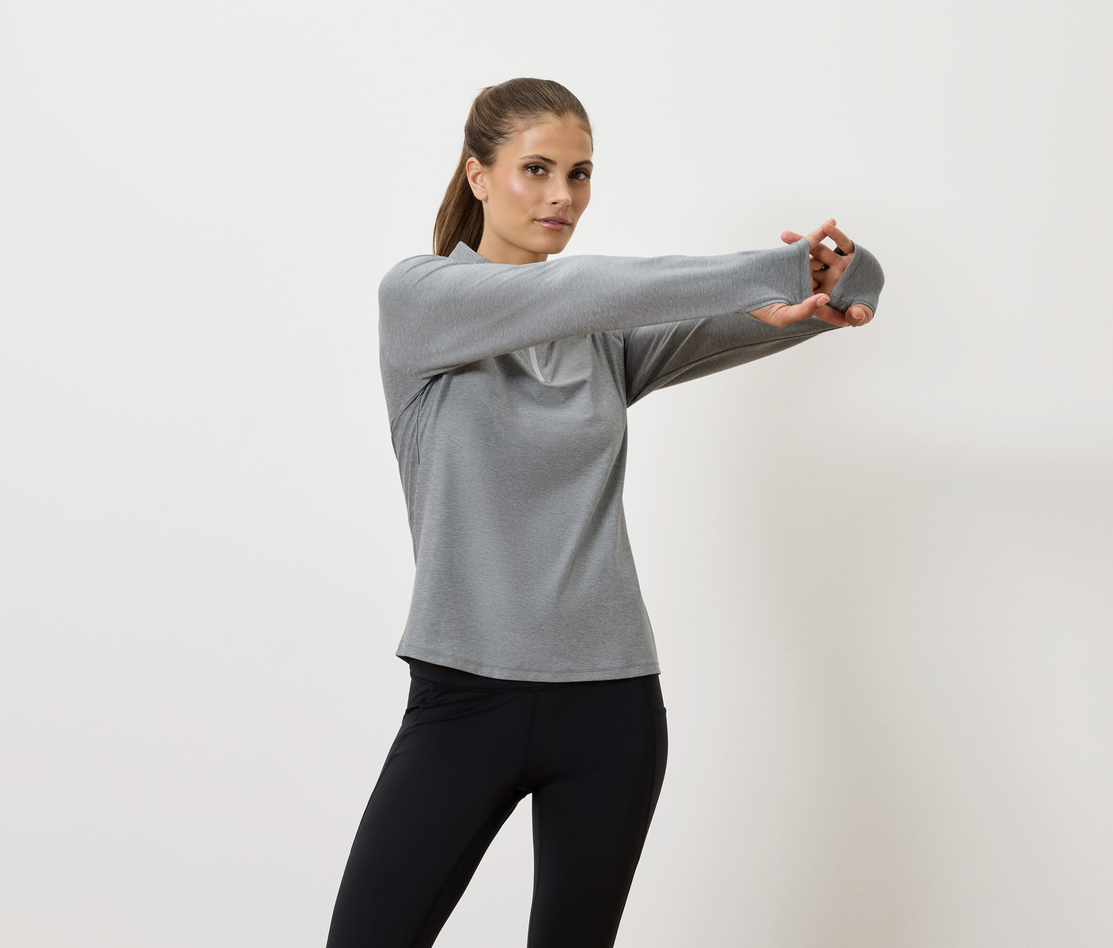 Žena si naťahuje ruky v šedej košeli s dlhým rukávom a čiernych legínach.
