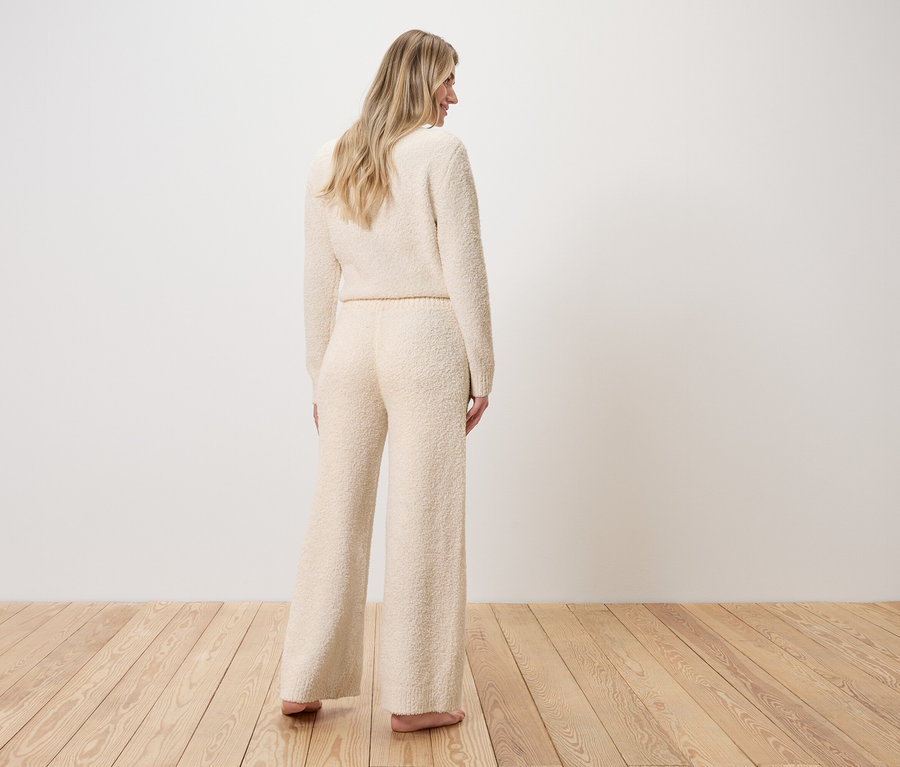 Žena stojí chrbtom, oblečená do svetra a širokých nohavíc z buklé úpletu.