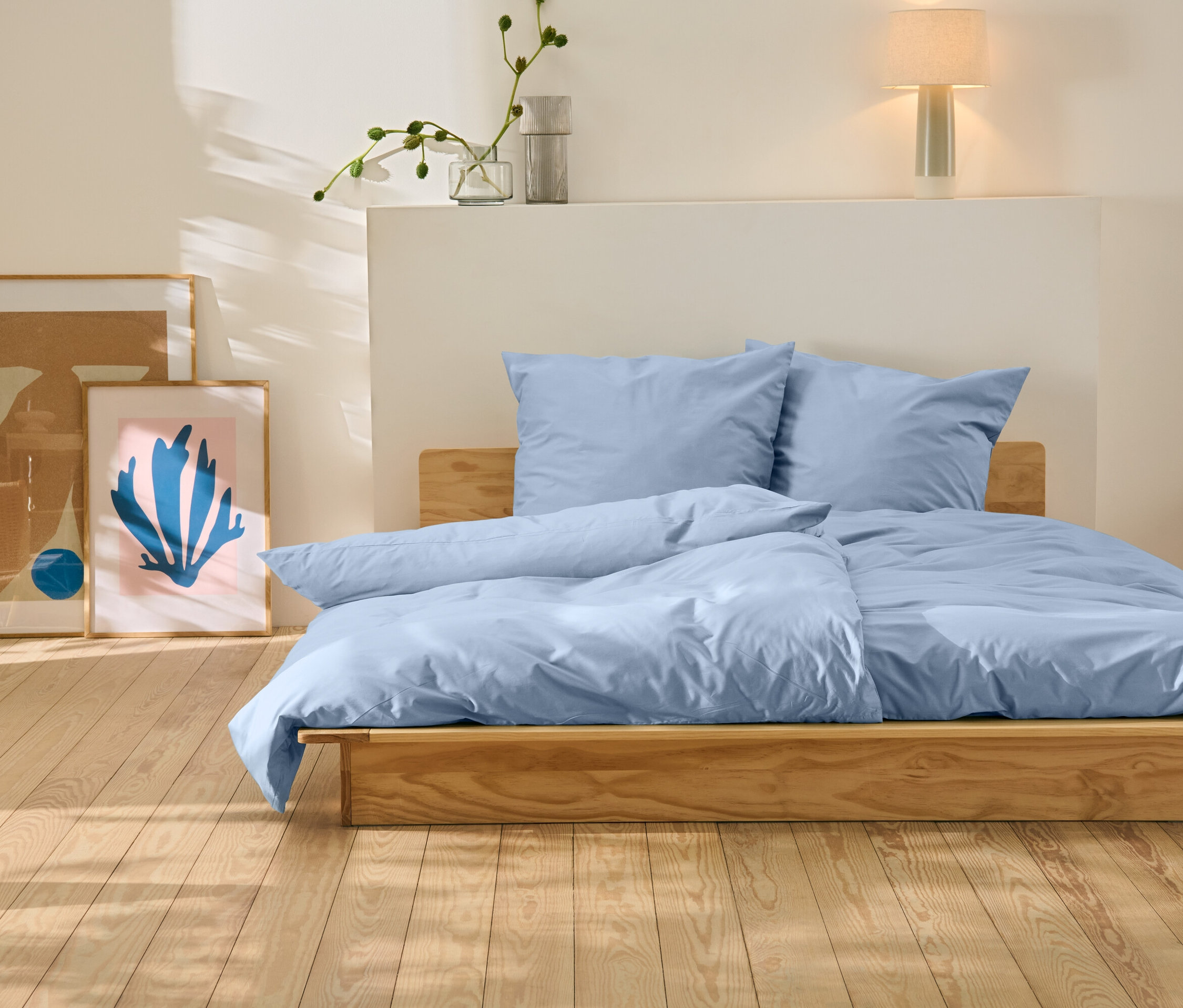 Posteľ s modrými obliečkami stojí na drevenej podlahe. Za posteľou sú dva obrazy.