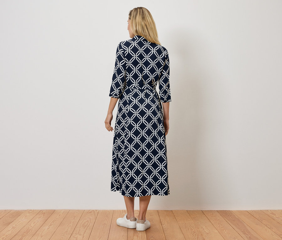 Pohľad zozadu na ženu v tmavomodrých šatách s bielym geometrickým vzorom a bielych teniskách.