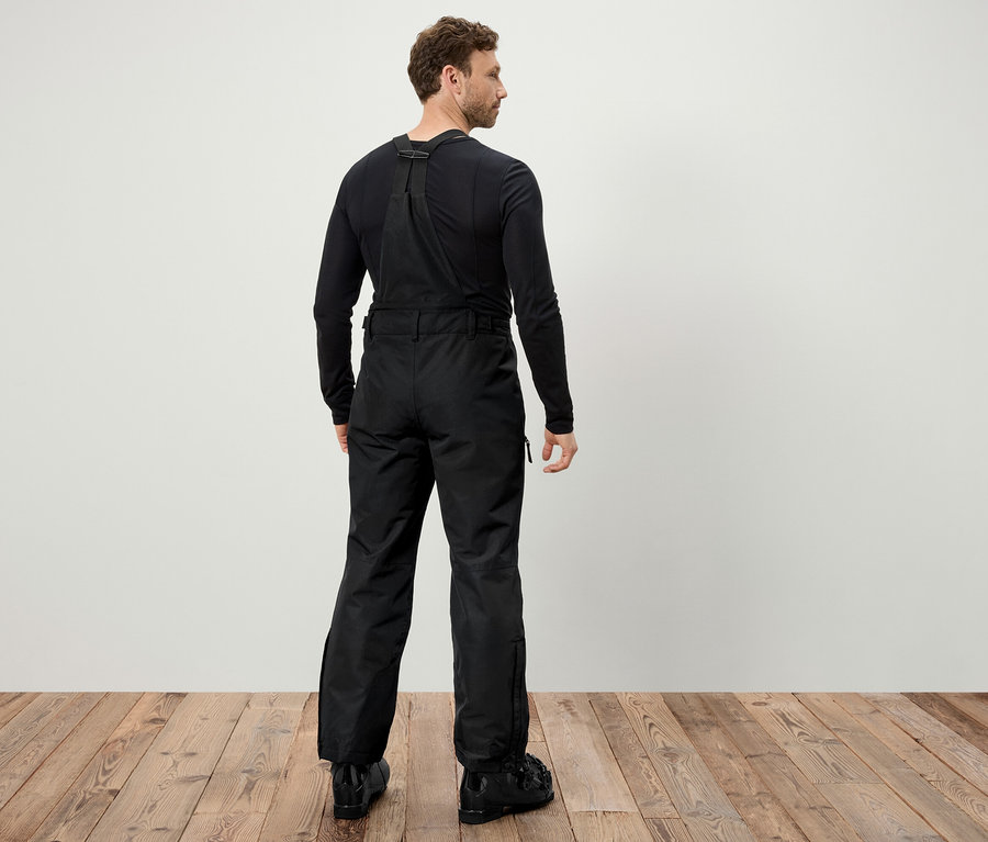 Muž chrbtom k fotoaparátu má na sebe čierne lyžiarske nohavice s trakmi a čierne lyžiarske topánky.