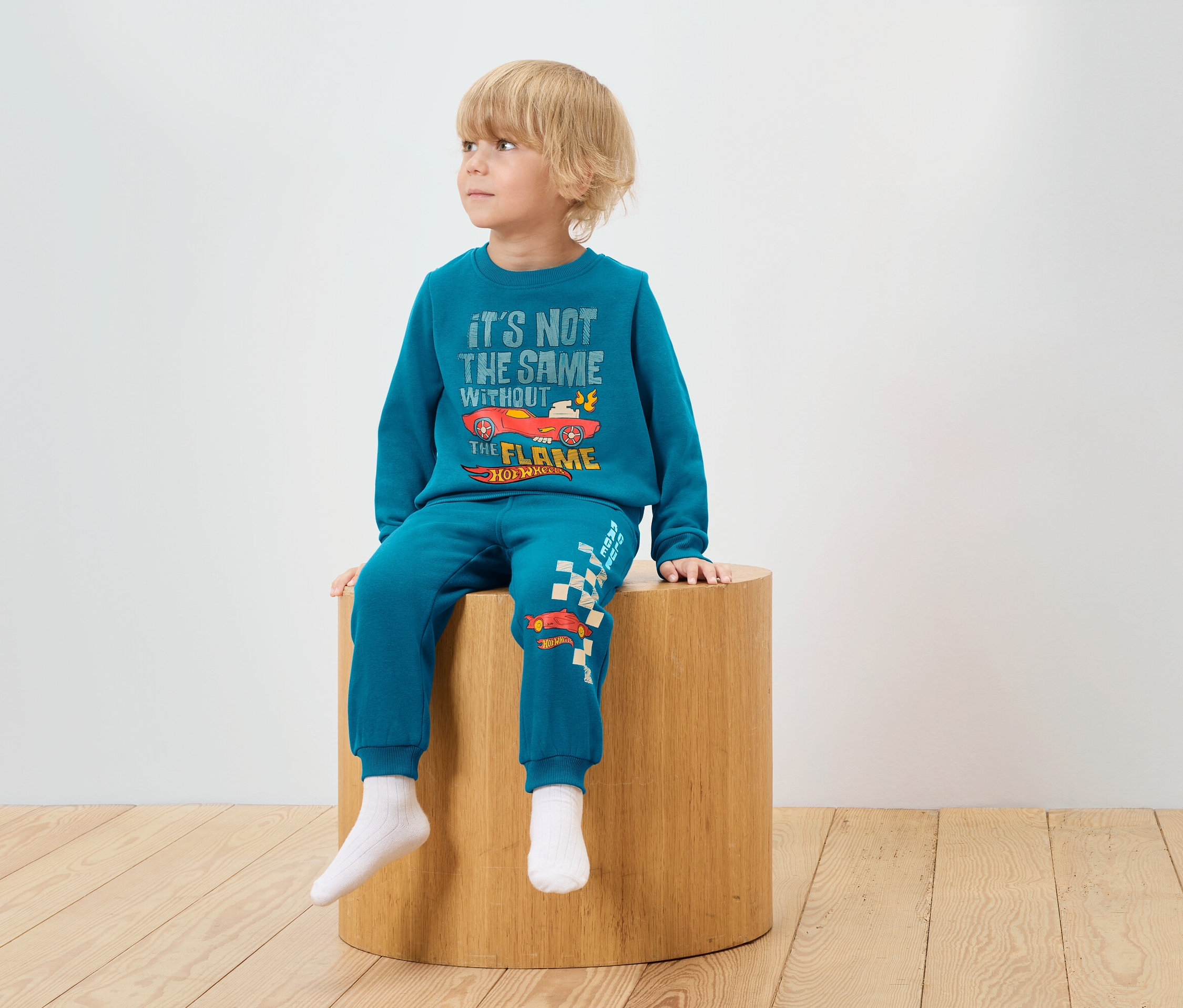 Chlapec sedí na drevenej stoličke a má na sebe modrú súpravu s potlačou Hot Wheels.