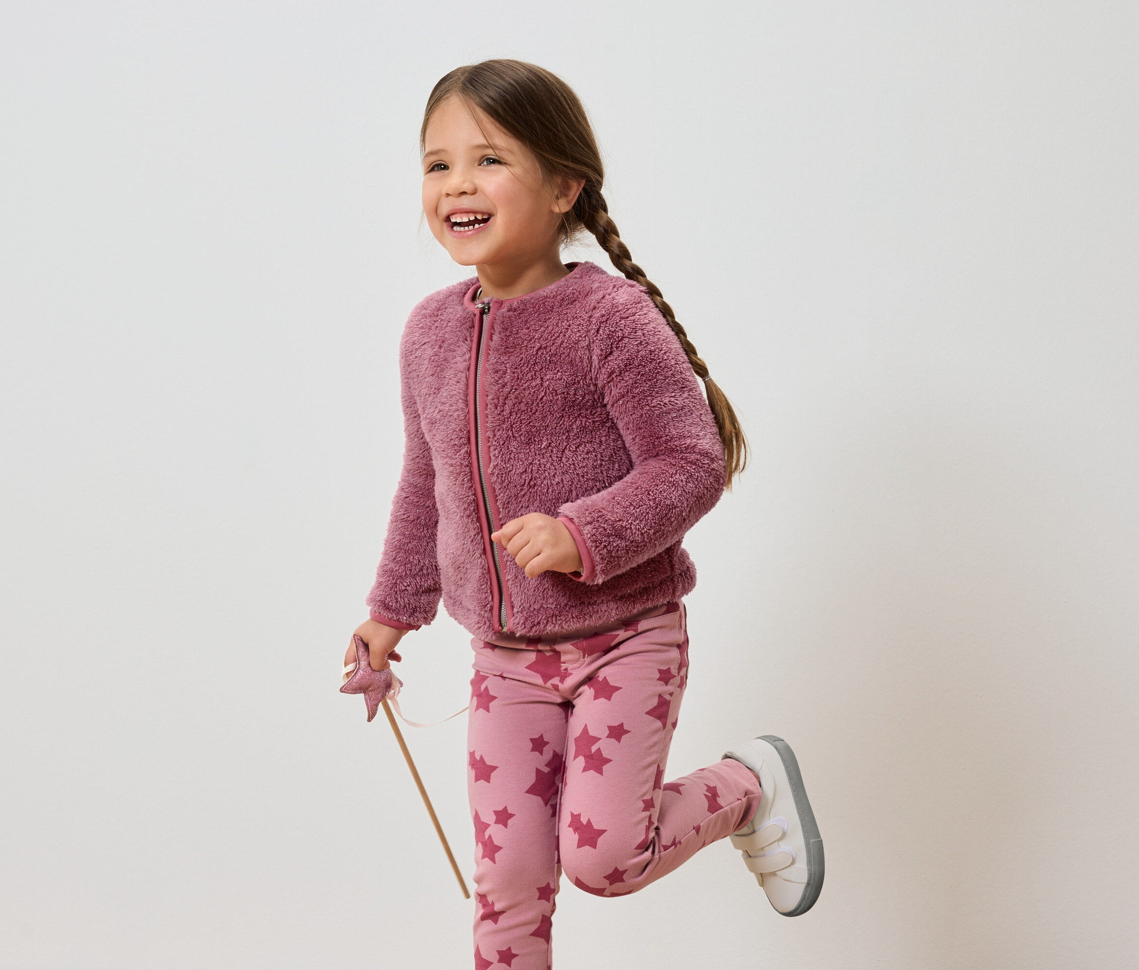 Usmievajúce sa dievča s hnedými vlasmi v ružovej bunde a nohaviciach s hviezdami drží prútik.