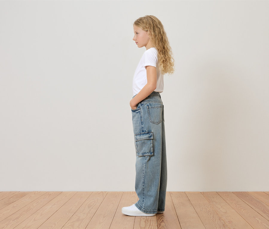 Mladé dievča s blond kučeravými vlasmi stojí bokom a má na sebe biele tričko, široké džínsy s bočným vreckom a biele tenisky.