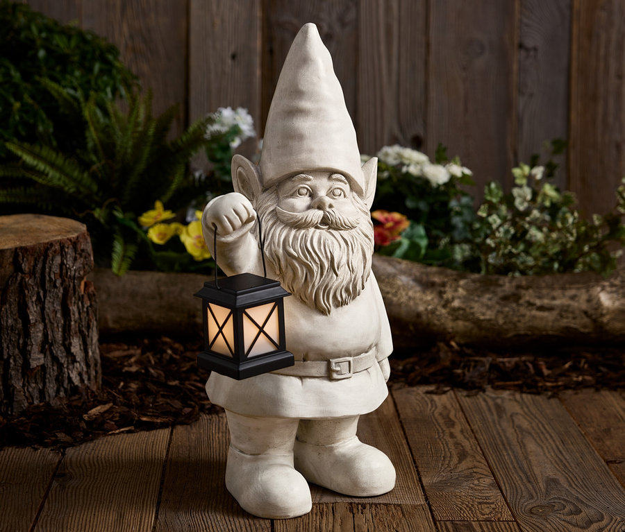 Kamenný záhradný trpaslík drží čiernu lampu pred dreveným pozadím.