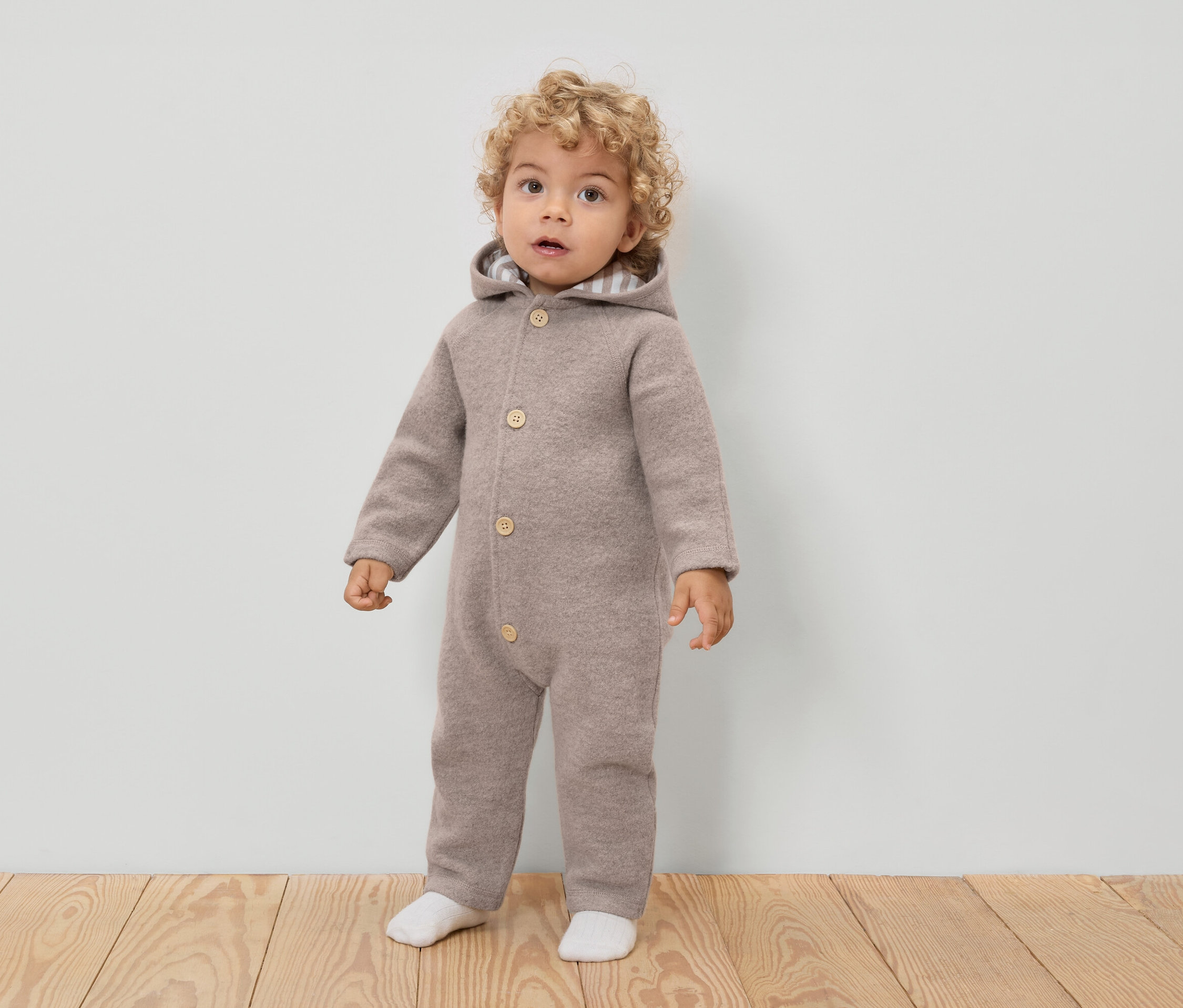 Malé dieťa s kučeravými vlasmi má na sebe svetlohnedú vlnenú kombinézu s kapucňou a gombíkmi a biele ponožky.