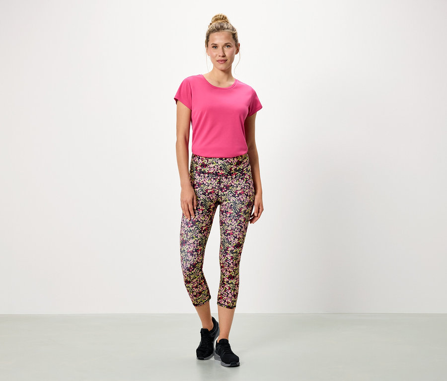 Žena stojí na bielom pozadí v ružovom tričku a kvetinových legínach.