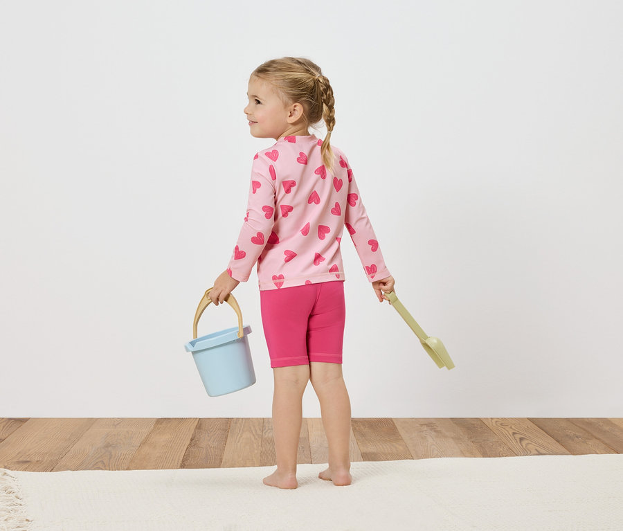 Dievča s blond vrkočmi, ružovou košeľou so srdiečkami, ružovými šortkami, svetlomodrým vedierkom a zelenou lopatkou.