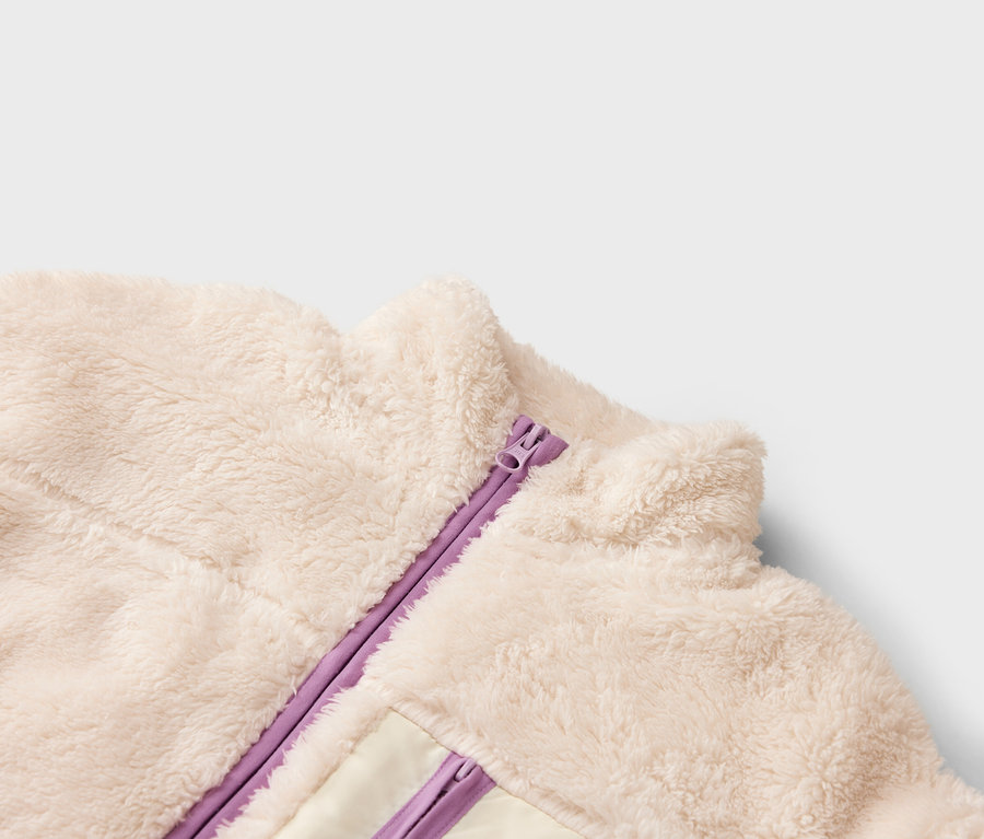 Detailný záber krémovej fleecovej bundy s fialovým zipsom.