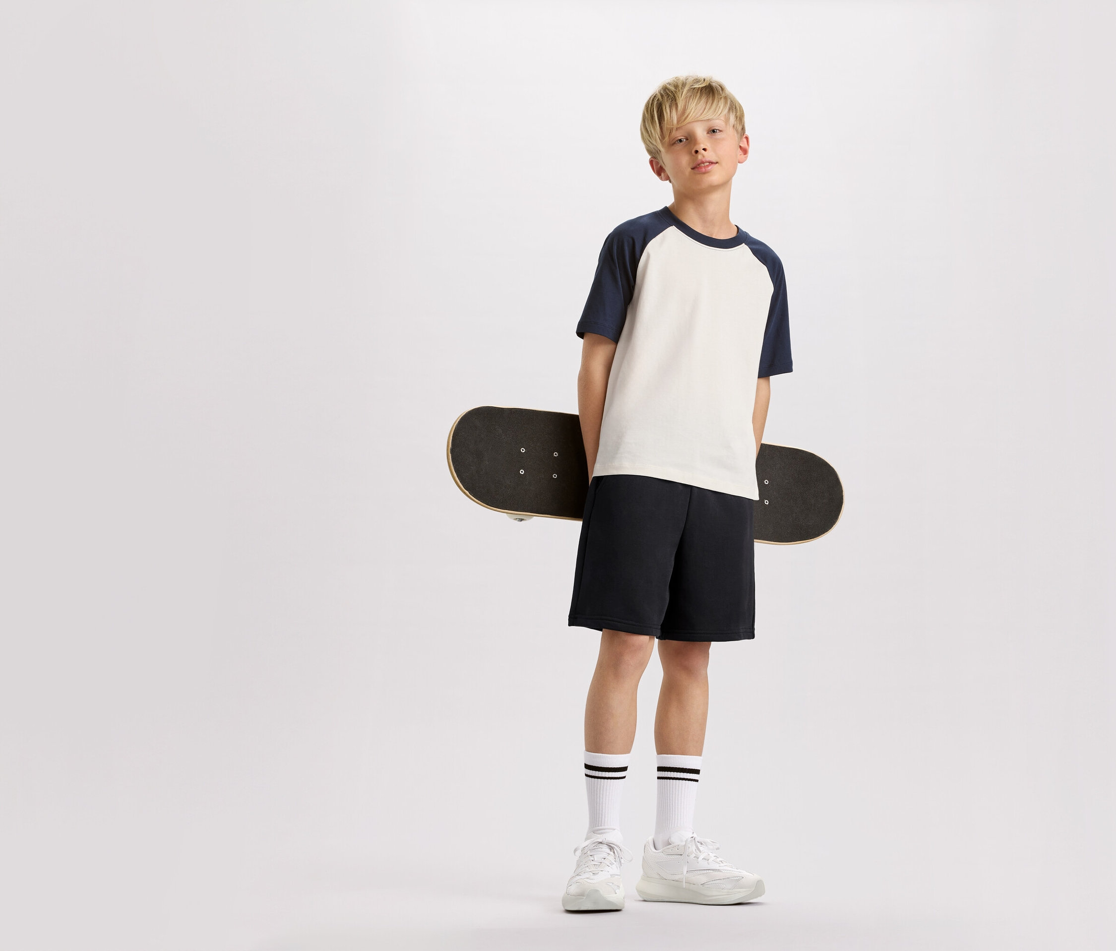 Blond chlapec drží za chrbtom skateboard, má na sebe tričko s tmavomodrými rukávmi, šortky a tenisky.