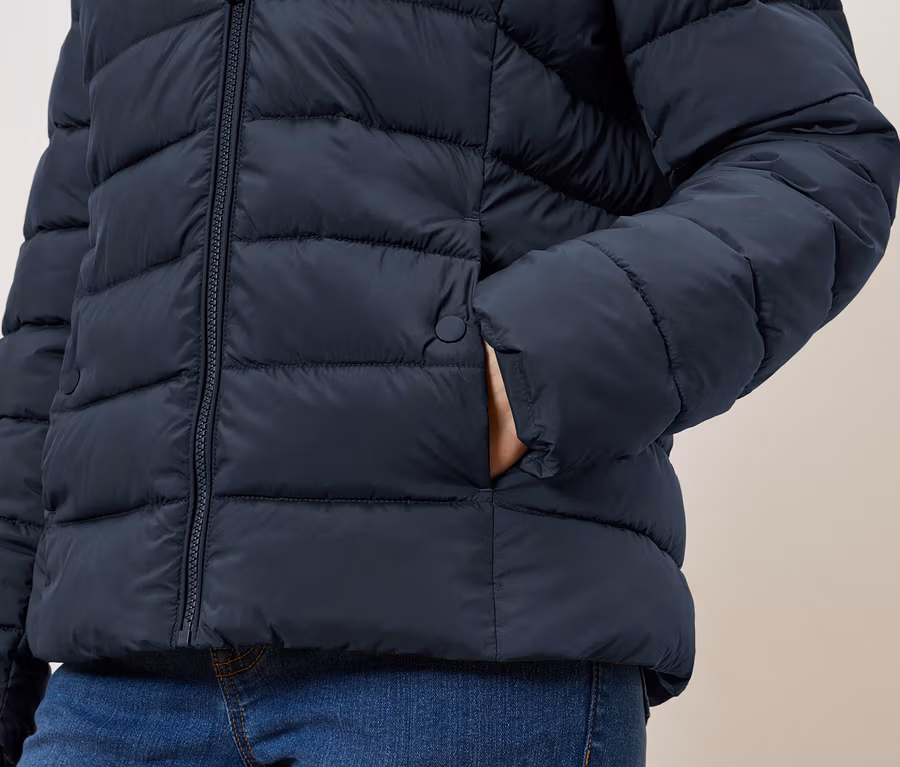 Detail osoby s rukou vo vrecku tmavomodrej prešívanej bundy na zips a modrých džínsov.