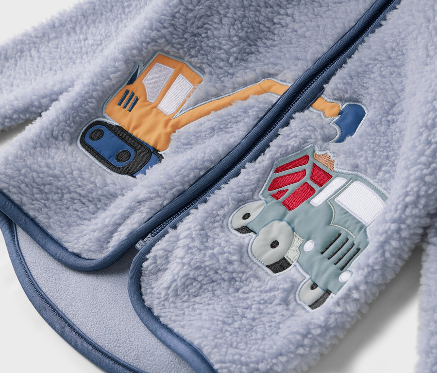 Detailný pohľad na svetlomodrú fleecovú bundu so zipsom a našitými motívmi bagra a nákladného auta.