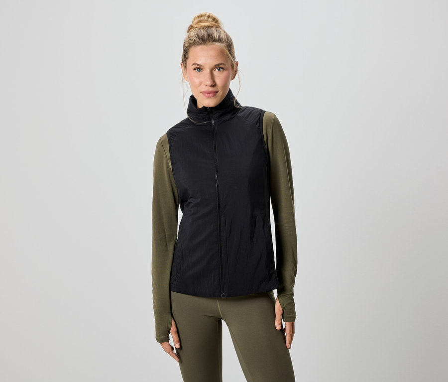 Žena s blond vlasmi má na sebe čiernu vestu cez zelené tričko s dlhým rukávom a legíny.