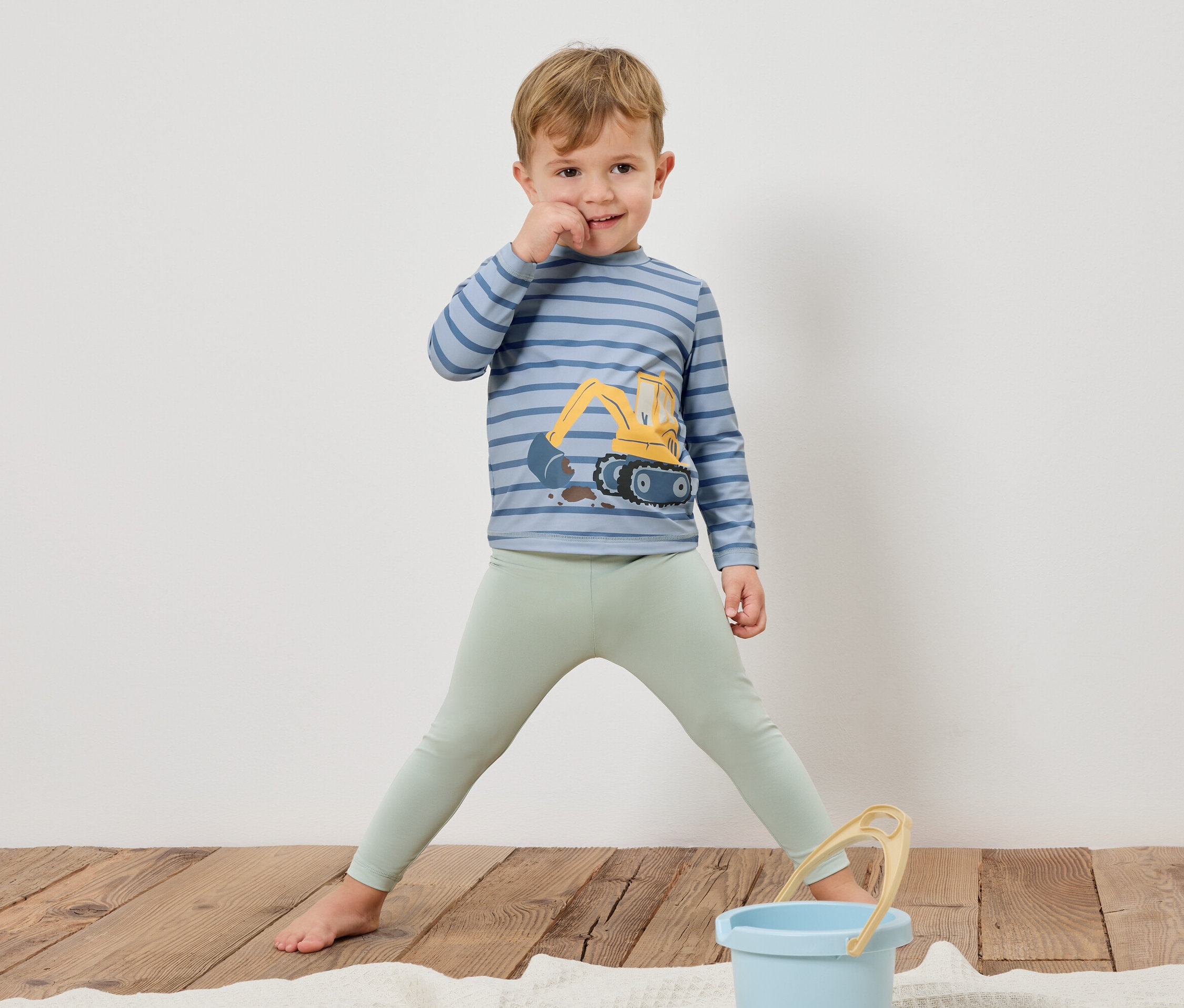 Chlapec stojí bosý na drevenej podlahe, má na sebe pruhované tričko s dlhým rukávom s bagrom a svetlozelené legíny. Pred ním stojí modré vedro.