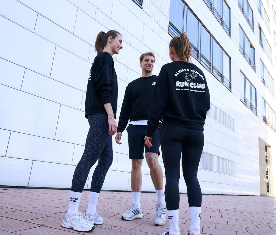 Traja ľudia, dve ženy a jeden muž, stoja usmiati a rozprávajú sa. Všetci majú na sebe čierne mikiny a biele unisex ponožky Coffee Crew. Muž má na sebe čierne športové šortky. Jedna žena má sivo-čierne športové legíny »ActivePower«, druhá čierne športové legíny »ActivePower«.