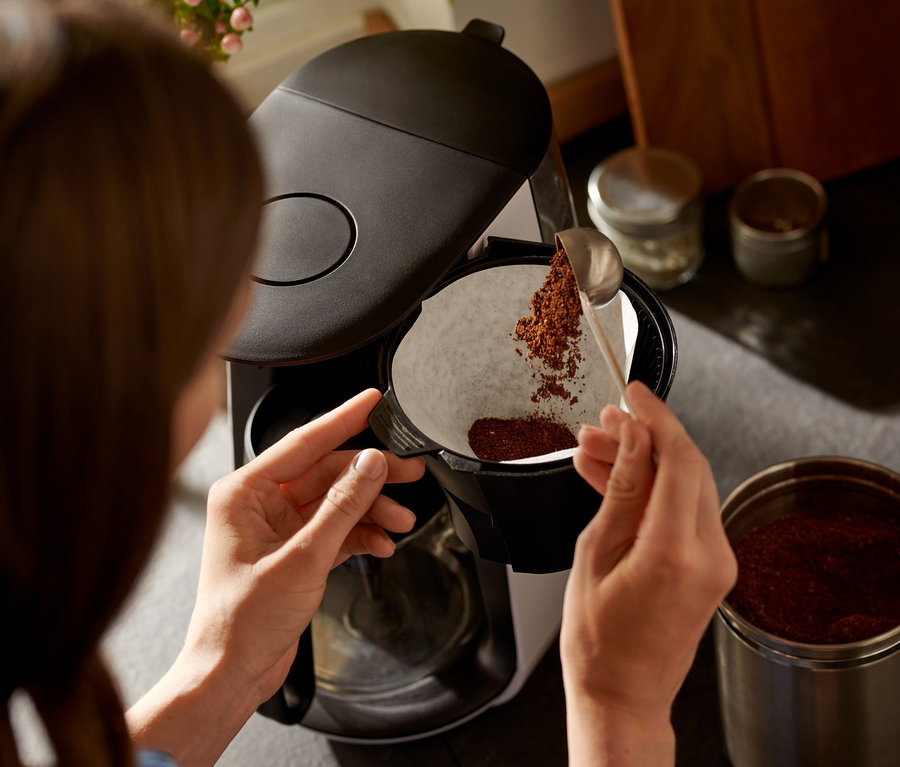 Žena sype kávu lyžicou do bieleho filtra kávovaru »Let's Brew«.