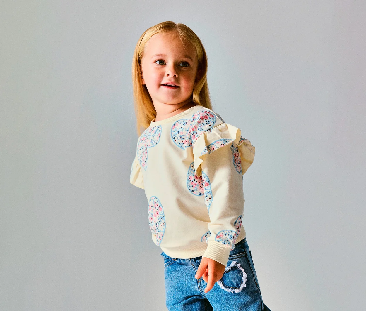 Mladé dievča s blond vlasmi má na sebe krémový NAME IT Kinder-Sweater s kvetinovými vzormi sŕdc a volánmi na ramenách, modré džínsy, pozerá doprava.