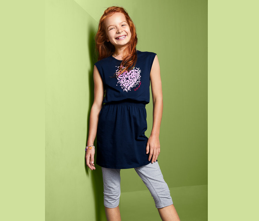 Usmievajúce sa dievča pózuje v tmavomodrých džersejových šatách s obojstrannými flitrami a sivých legínach.