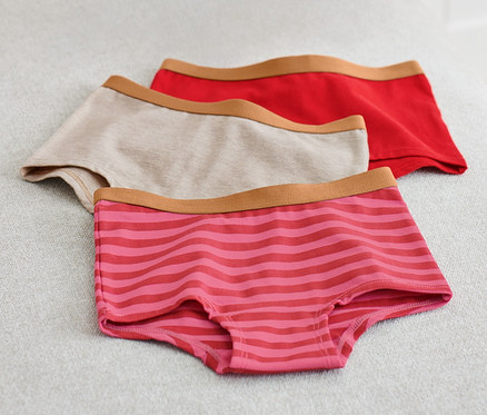 Na svetlosivom povrchu ležia troje detské nohavičky, jedny béžové, jedny červené a jedny ružové s prúžkami. Vľavo je ružová deka.