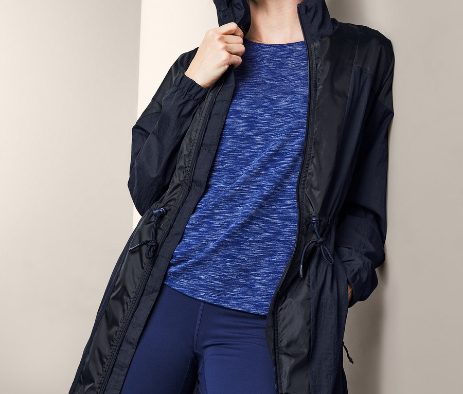 Blondínka opretá o stenu má na sebe funkčnú bundu, modré tričko a legíny.