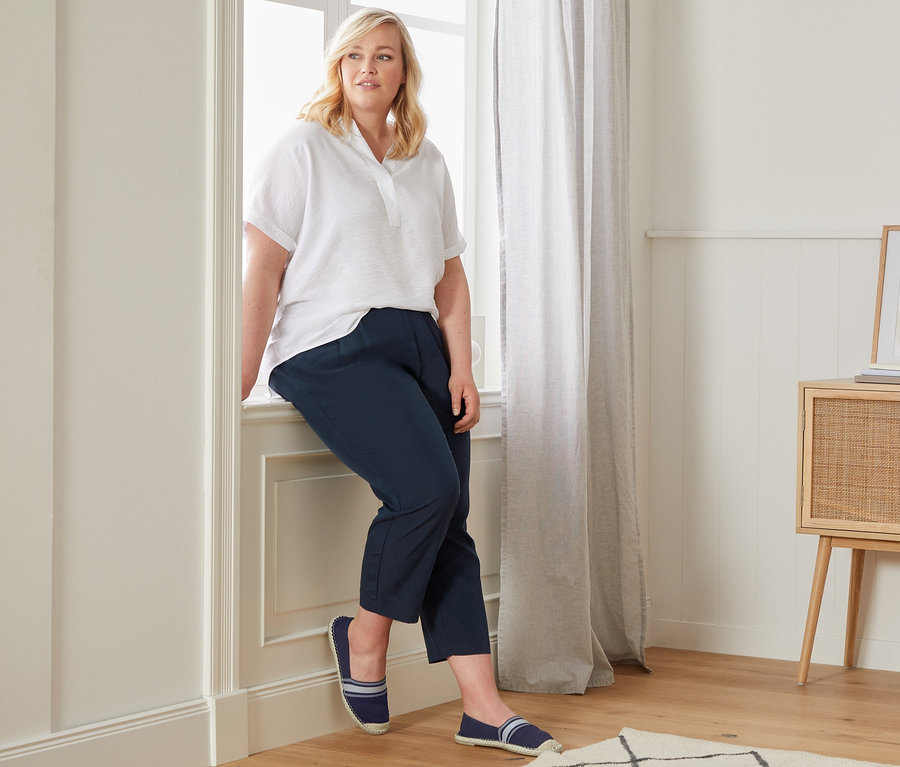 Žena sedí na parapete v bielej blúzke v oversize štýle, ležérnych nohaviciach chino a espadrilkách.