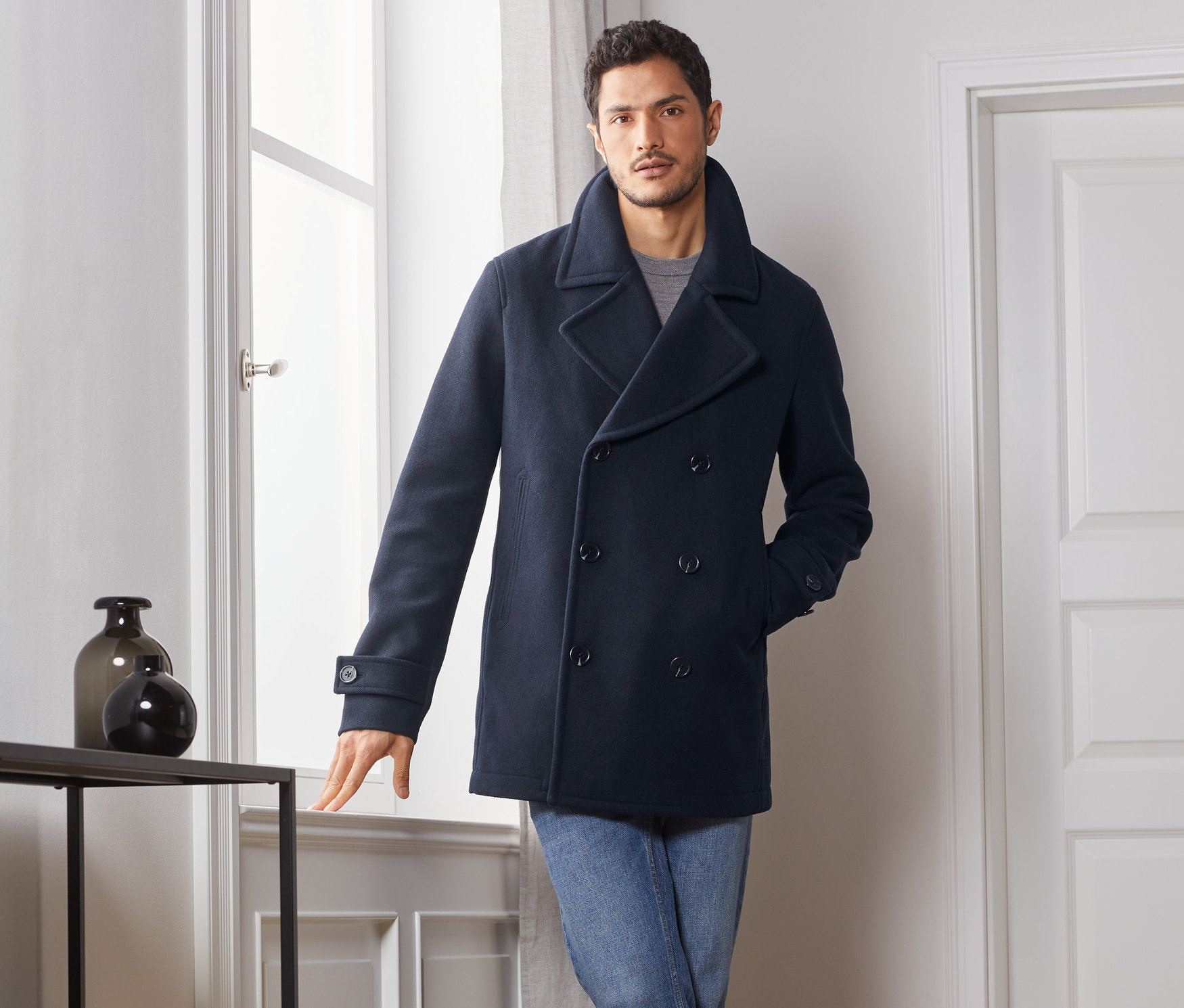 Muž pózuje pri okne v krátkom kabáte, džínsoch a topánkach.