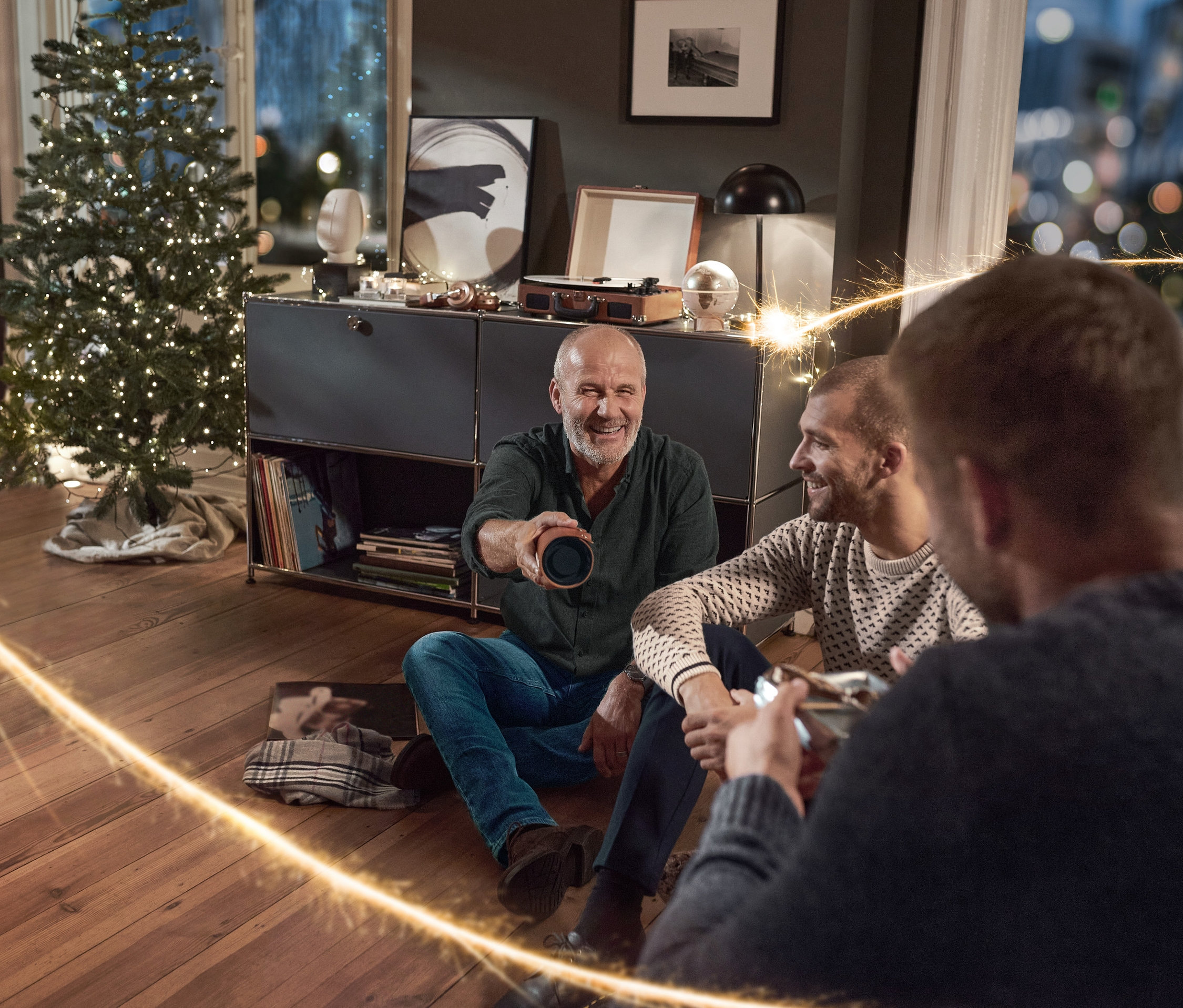 Traja muži sedia na podlahe v obývacej izbe, jeden drží Vintage reproduktor s Bluetooth® a obalom z koženky. Majú na sebe Džínsy, Pletený pulóver a Flanelovú košeľu.