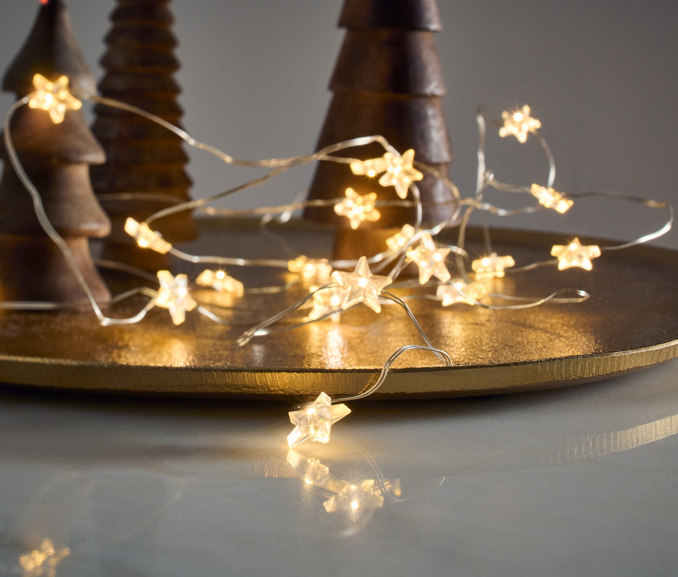 Svetelná reťaz s mikro LED, hviezdy, leží na zlatom podnose vedľa drevených ozdobných stromčekov.