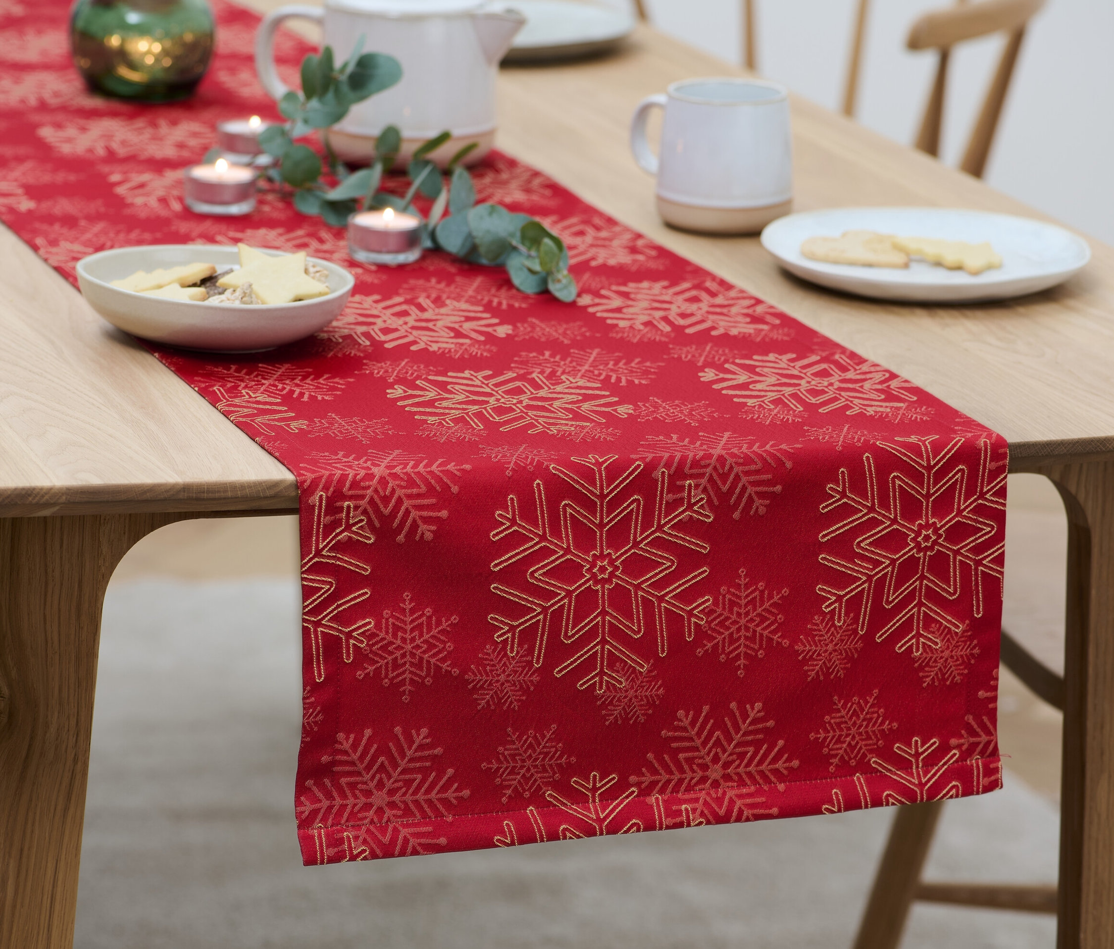Na drevenom stole je červený žakárový behúň so vzorom snehových vločiek, na ktorom sú taniere s koláčikmi, sviečky a kanvica.