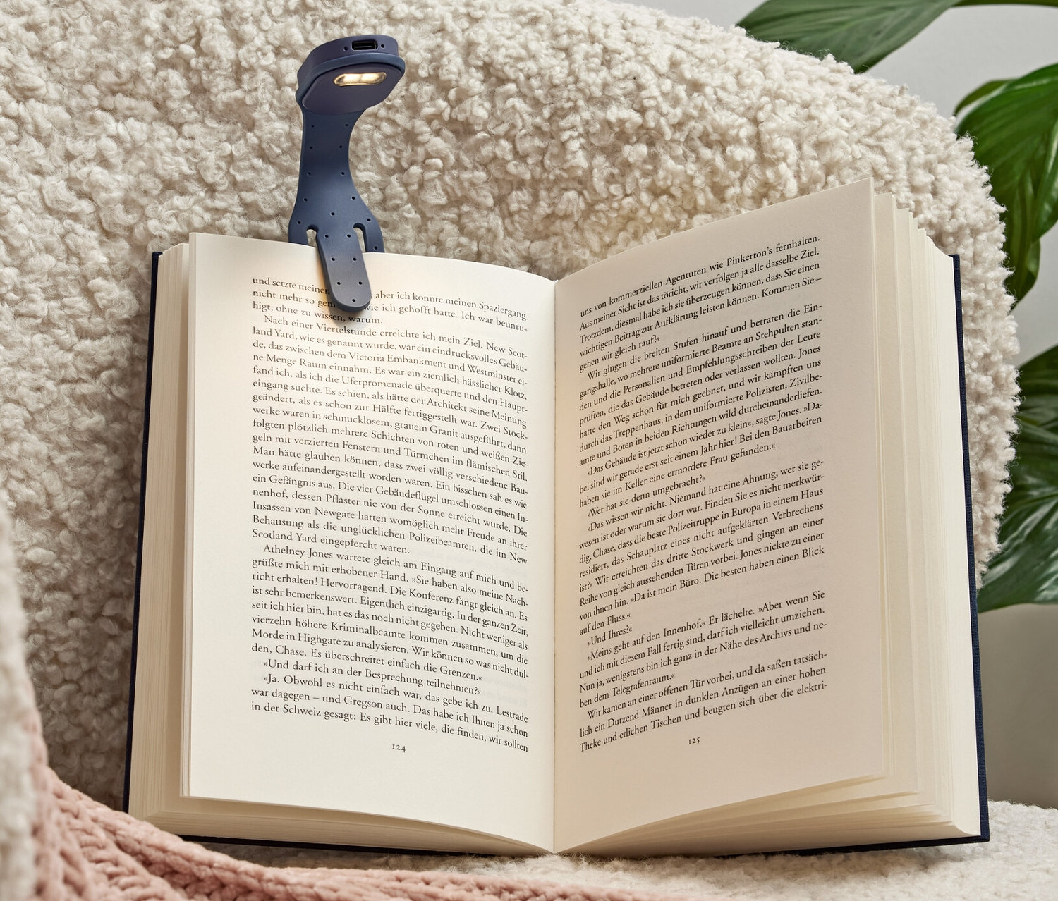 Modrá mini LED lampa na čítanie s akumulátorom pripnutá na otvorenej knihe ležiacej na kresle.