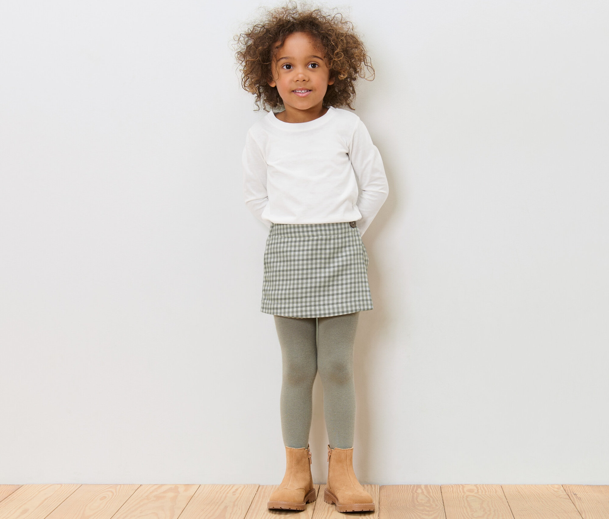 Dievča stojí a má na sebe detskú nohavicovú sukňu, biele tričko a hnedé topánky.