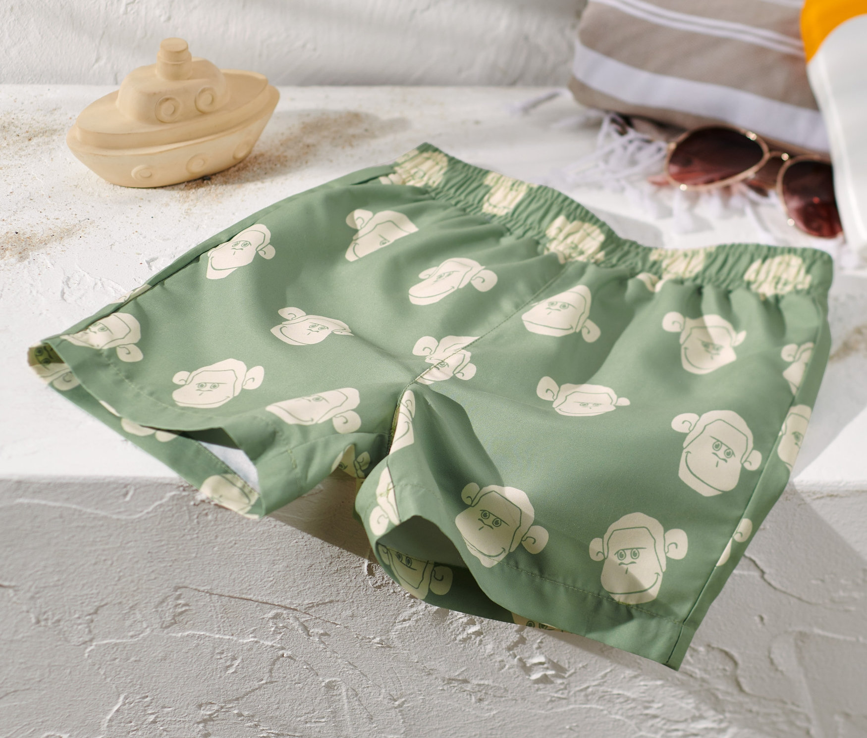 Zelené detské šortky na kúpanie s potlačou opíc ležia na bielom povrchu vedľa hračky lode, uteráka, slnečných okuliarov a opaľovacieho krému.