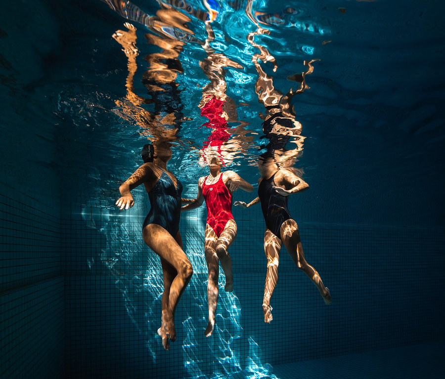 Tri ženy plávajú pod vodou v bazéne. Jedna z nich má na sebe arena Shapewear Damen Lara C-Cup Badeanzug.