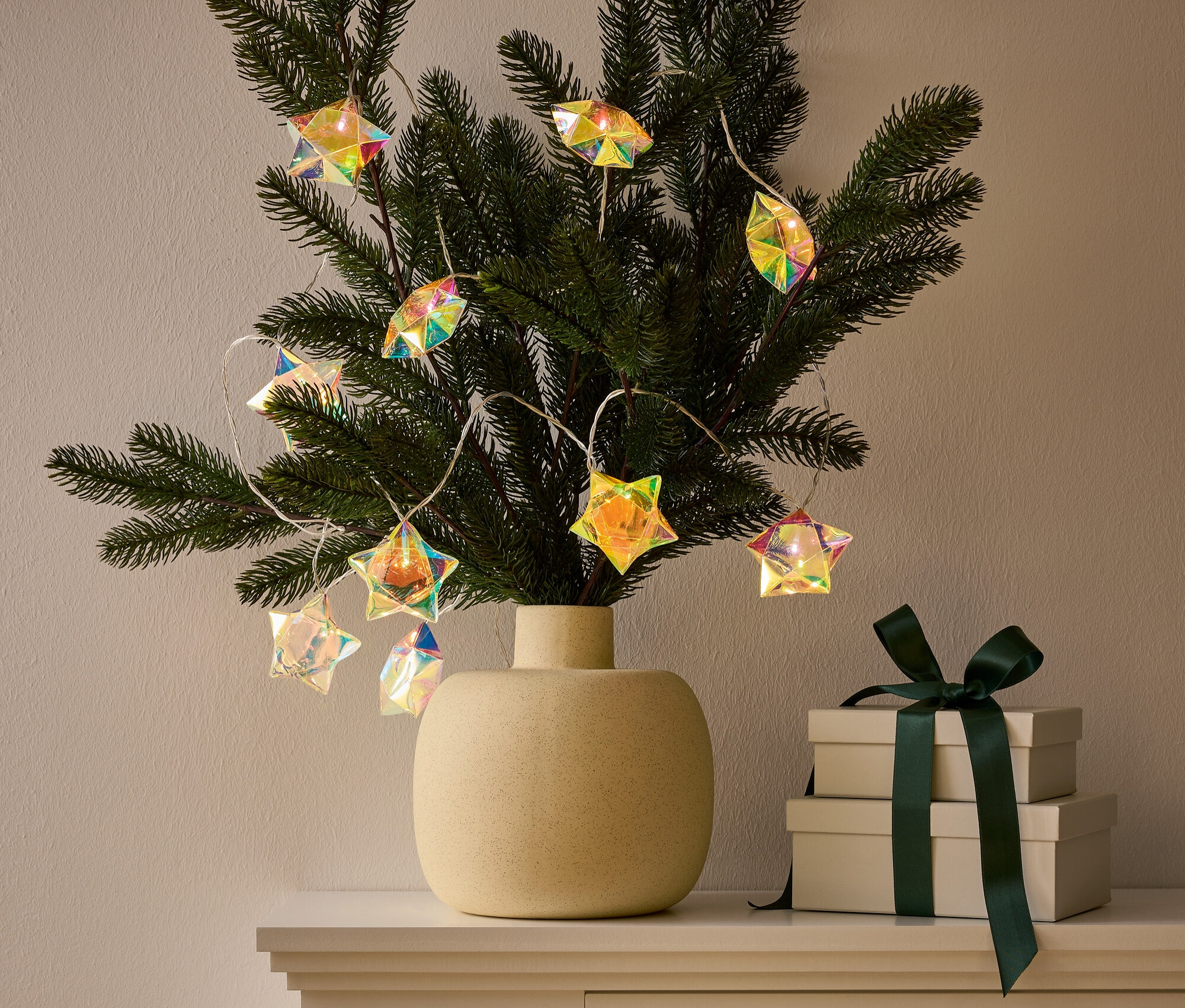 Váza s vetvičkou zdobenou svetelnou reťazou v tvare hviezdy stojí na skrinke vedľa dvoch darčekových krabíc s tmavozelenou stuhou.