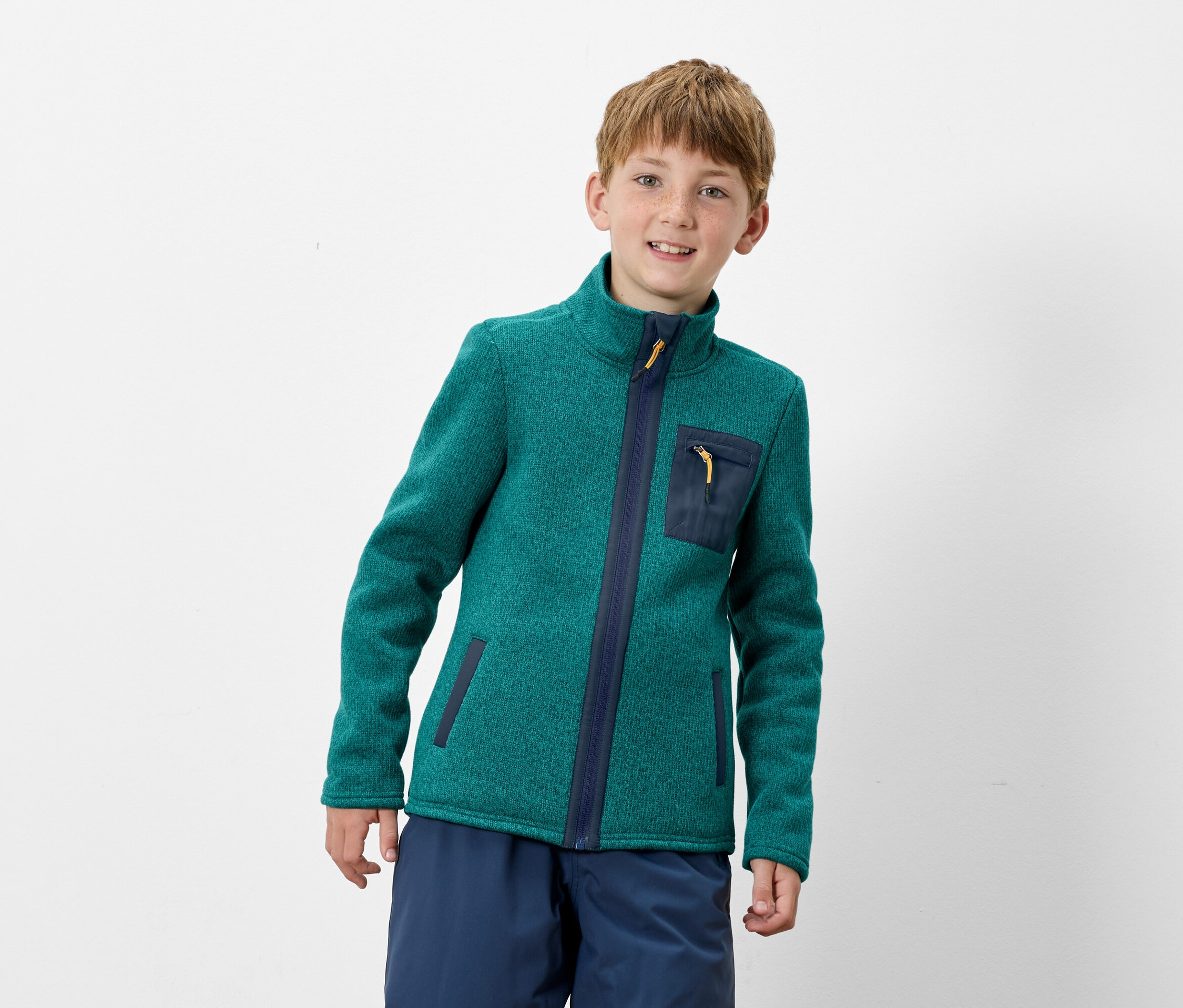 Chlapec má na sebe zelenú fleecovú bundu na zips s tmavomodrými detailmi a modré nohavice.