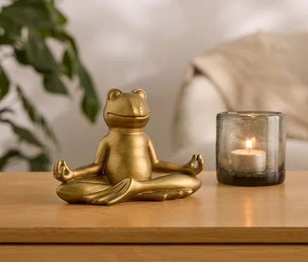 Zlatá soška žaby sedí v meditačnej pozícii vedľa sklenenej nádoby so zapálenou sviečkou.