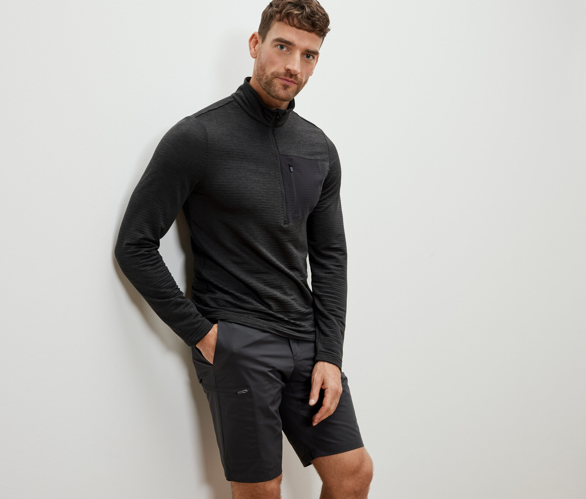 Muž opretý o stenu má na sebe antracitové funkčné outdoorové tričko s dlhým rukávom a šortky.