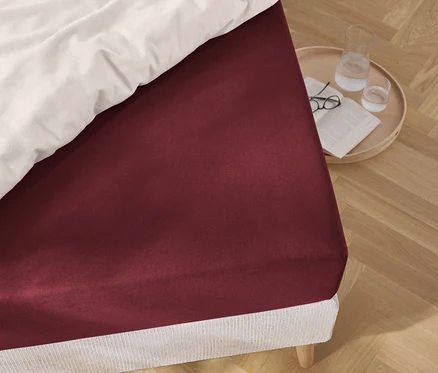 Detail červenej plachty na posteli, vedľa ktorej stojí okrúhly stolík s okuliarmi, knihou a pohárom vody.