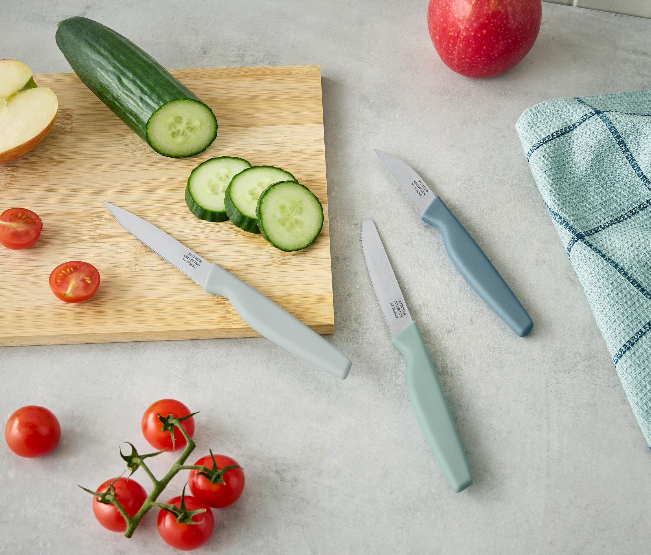 Na drevenej doske leží polovica jablka, uhorka a nakrájané plátky uhorky. Vedľa ležia tri nože a cherry paradajky.