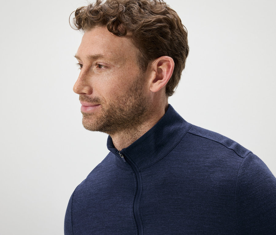 Portrét muža s hnedými vlnitými vlasmi a bradou, oblečeného v modrom svetri na zips.